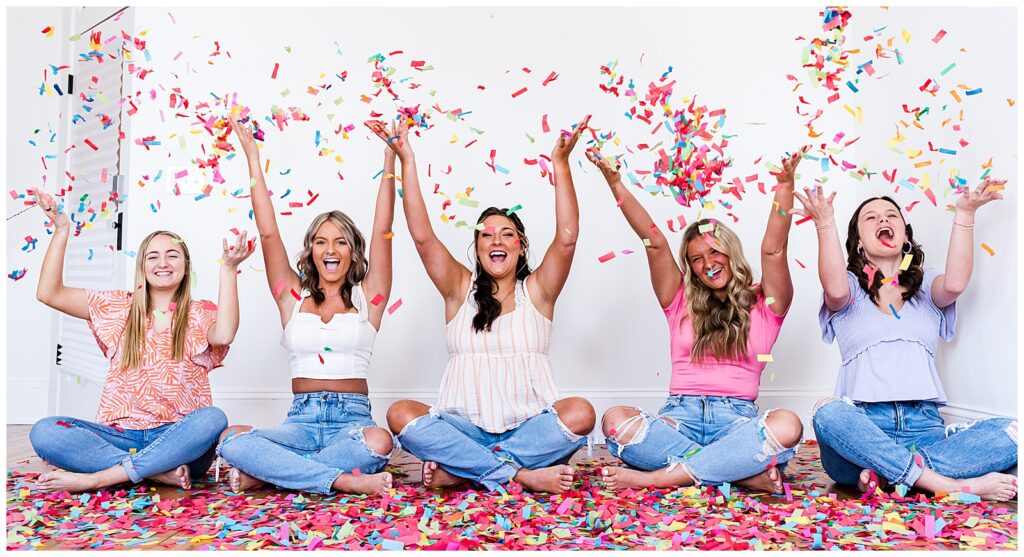 High school senior spokesmodels sit on the floor laughing as they throw confetti up in the air for photos. 
