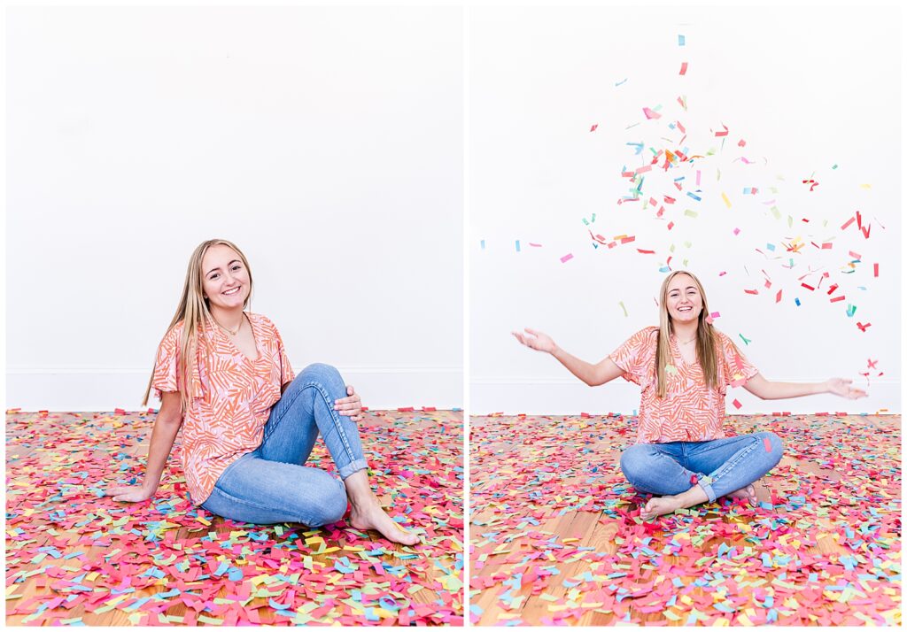 A high school senior spokesmodel wearing jeans and a pink and orange flowered top sits on the floor laughing as she throws confetti up in the air for pictures. 
