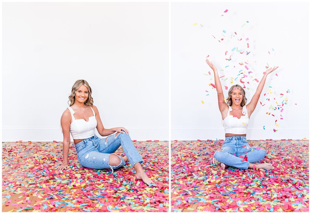 A high school senior spokesmodel wearing jeans and a white crop top sits on the floor laughing as she throws confetti up in the air for photos. 
