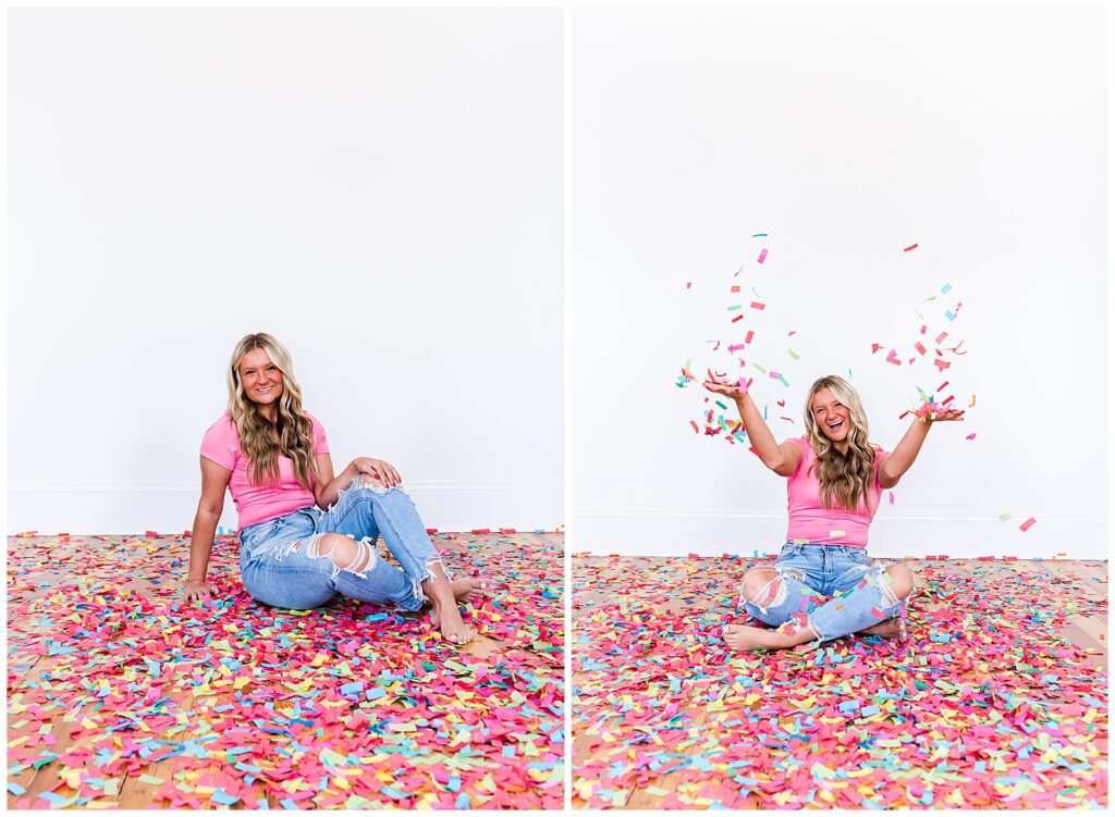 A high school senior spokesmodel wearing jeans and a pink top sits on the floor laughing as she throws confetti up in the air for photos. 