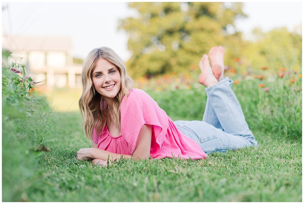 Senior girl wearing a pink top and jeans, laying in a field of wildflowers.
