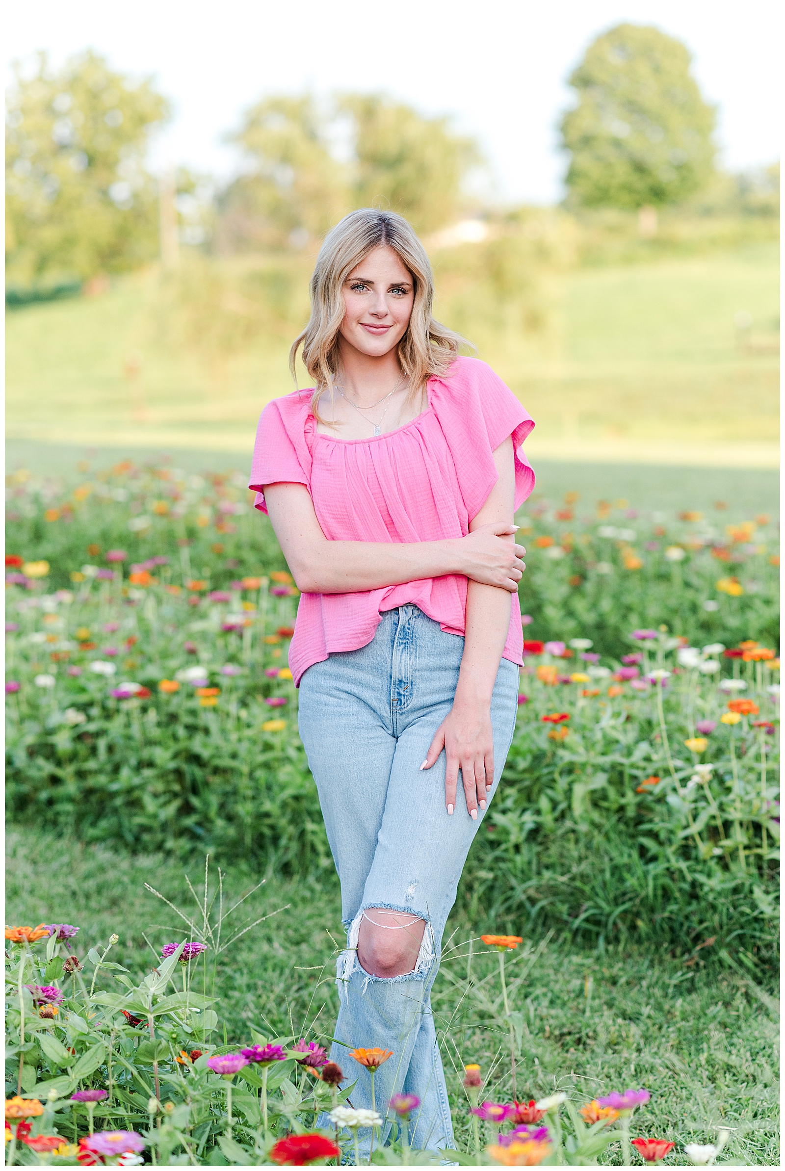Senior girl wearing a pink top and jeans, standing in a field of wildflowers.