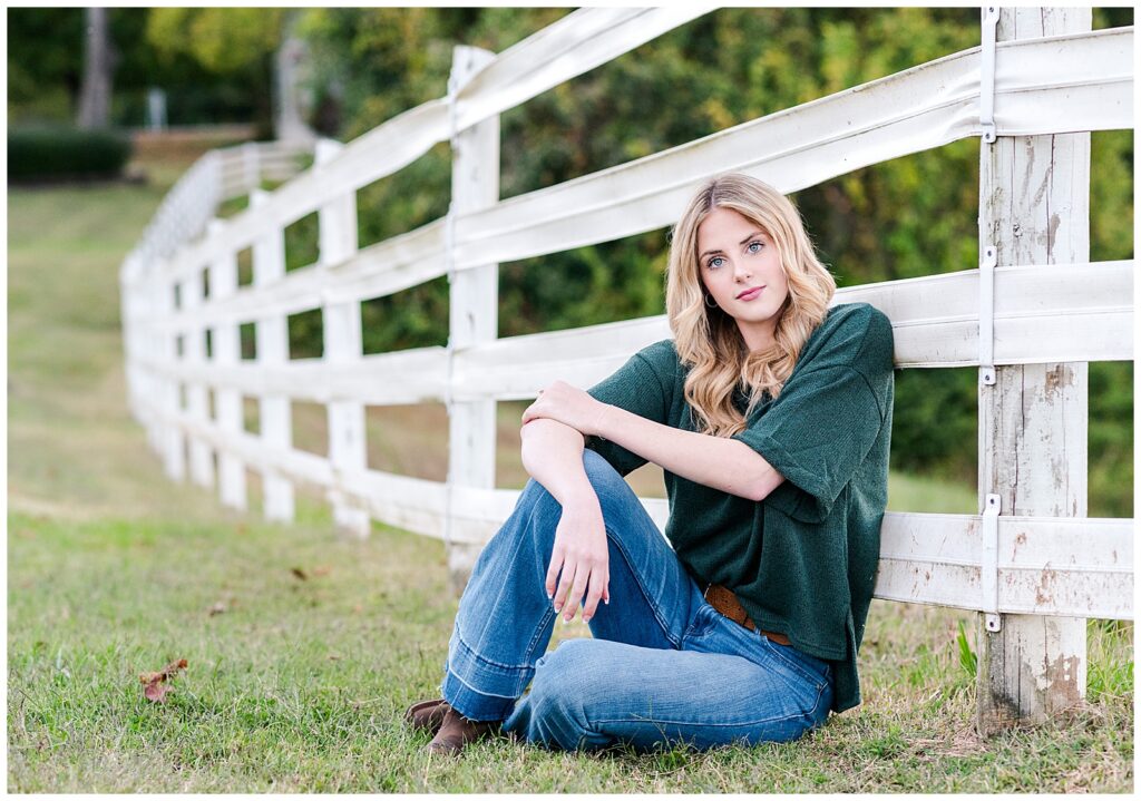 High school senior girl sitting down, leaning on a fence at Ellington Agricultural Center.