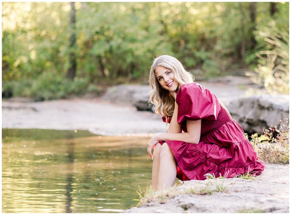 High school senior girl wearing a cranberry-colored dress, sitting on the creek bank rocks at Ellington Agricultural Center.
