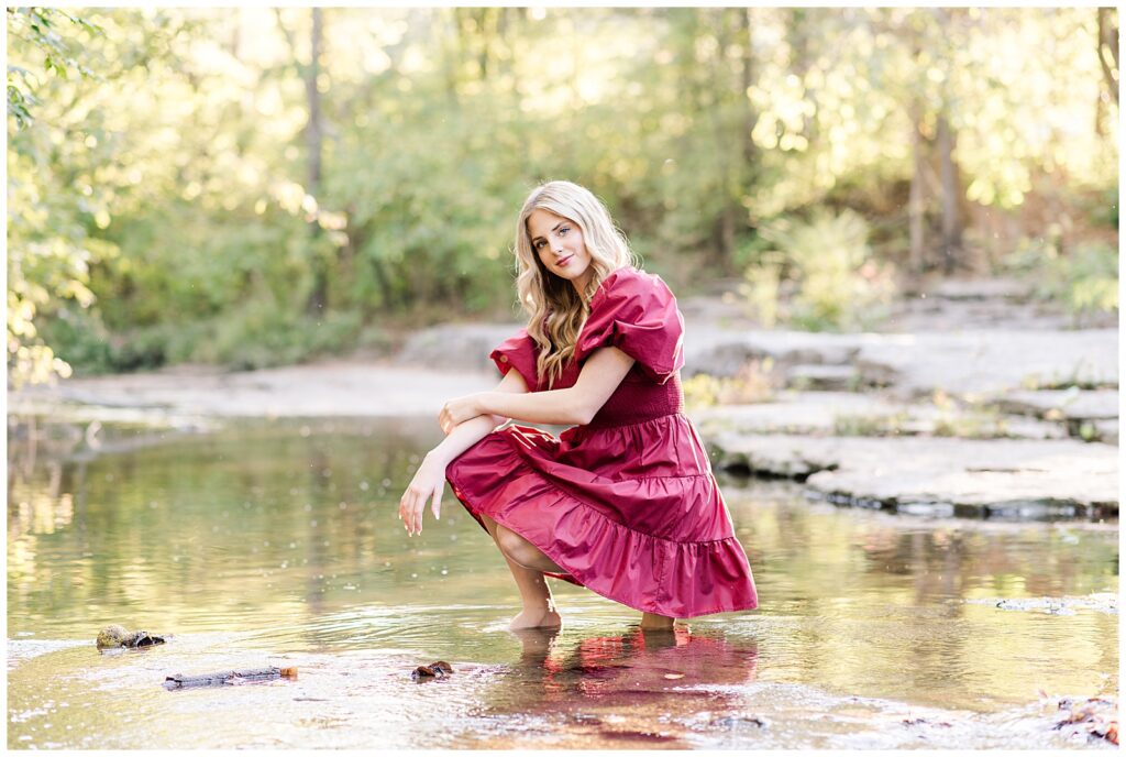 High school senior girl wearing a cranberry-colored dress, kneeling in the creek, at Ellington Agricultural Center.