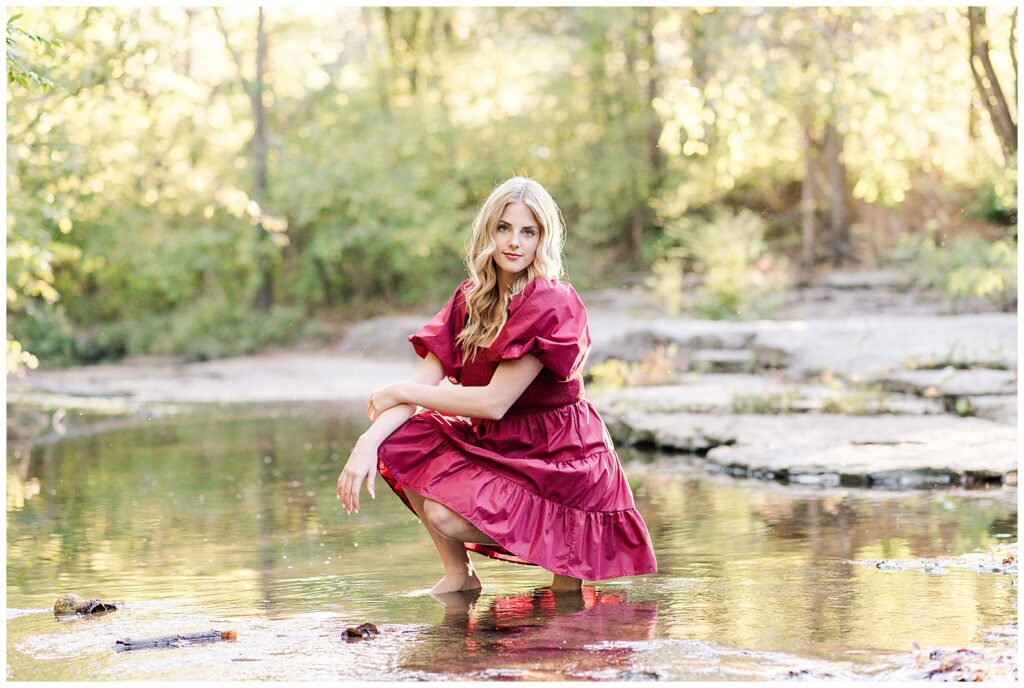 High school senior girl wearing a cranberry-colored dress, kneeling in the creek at Ellington Agricultural Center.