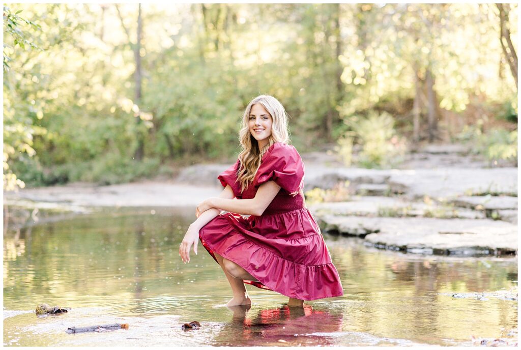 High school senior girl wearing a cranberry-colored dress, kneeling in the creek at Ellington Agricultural Center.