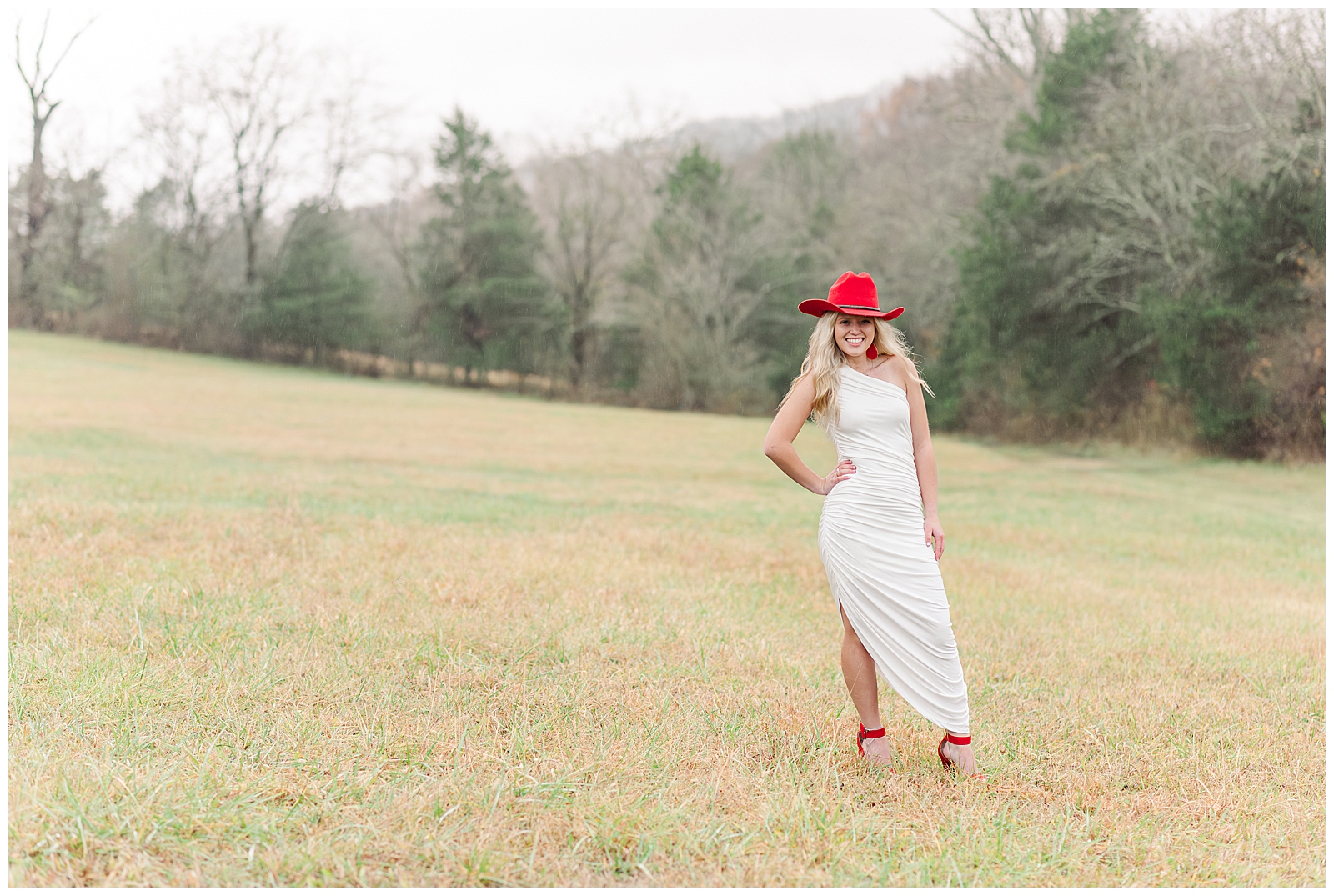 High school senior girl wearing a white dress, a red cowboy hat, and red shoes, standing in an open field for her private farm and lake senior session.