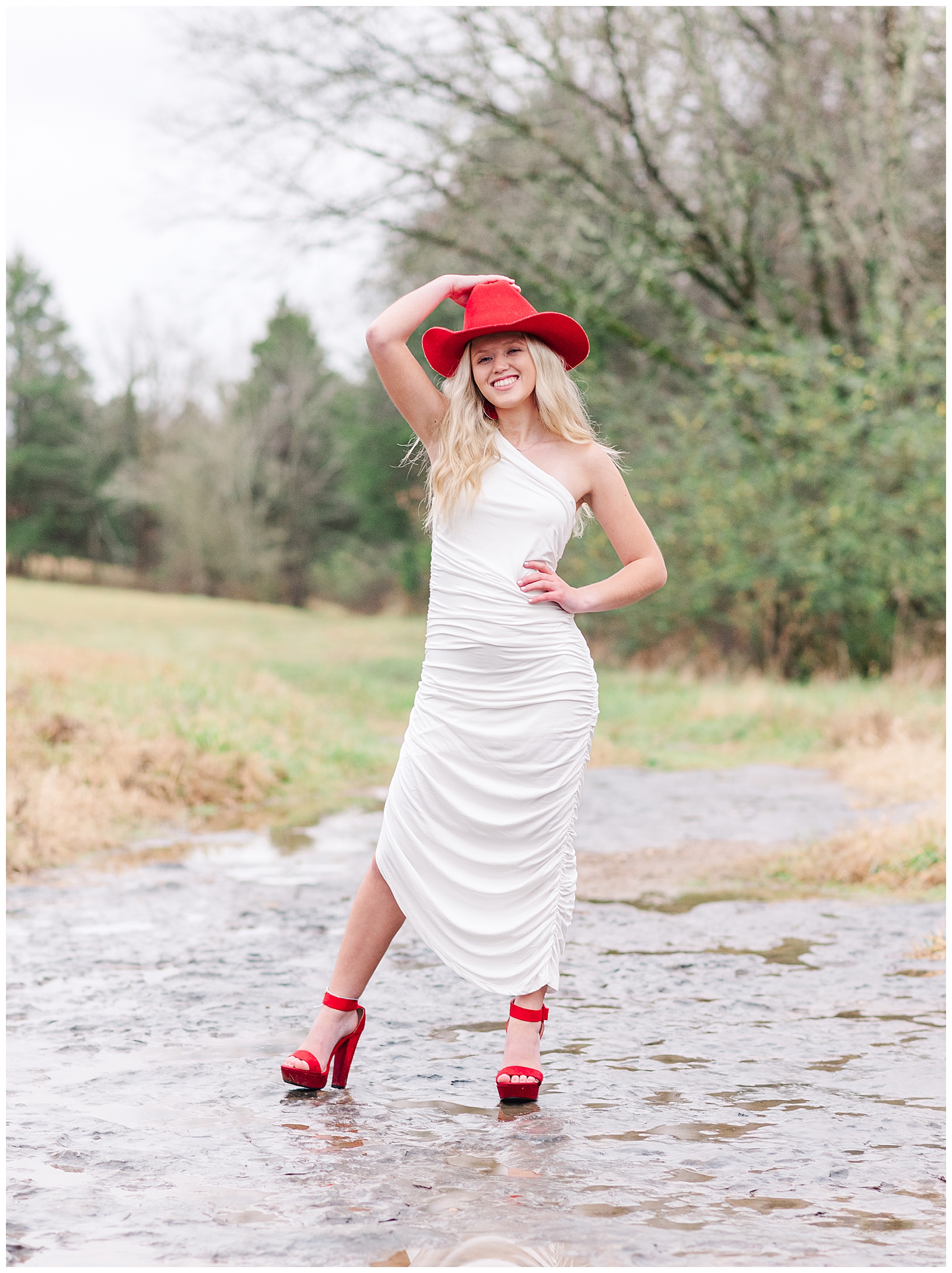 High school senior girl wearing a white dress, a red cowboy hat, and red shoes, standing in a dry creek bed for her private farm and lake senior session.