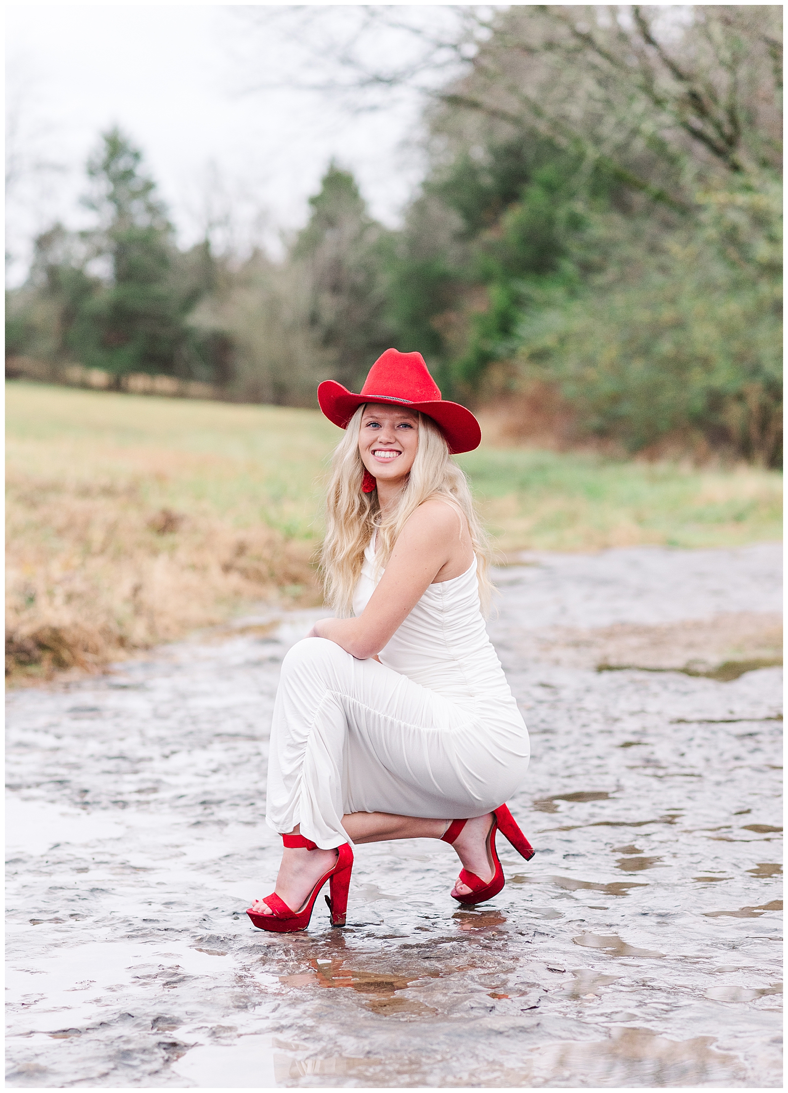 High school senior girl wearing a white dress, a red cowboy hat, and red shoes, kneeling in a dry creek bed for her private farm and lake senior session.